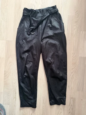 Svarta byxor i läderimitation från Boohoo. 42/14 - Svarta byxor i läderimitation från Boohoo. Hög midja med bälte och lös, bekväm passform. Mjukt material och snygg modell. Perfekta till både vardag och fest.  
