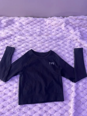 Svart ribbad långärmad topp NA-KD - Snygg svart ribbad långärmad topp från NA-KD med diskret logga på bröstet. Toppen har en croppad passform och rund halsringning, perfekt att styla med höga jeans eller kjol. Även träningsmaterial.