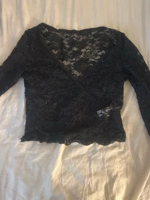 Svart spetsblus med v-ringning - Snygg svart blus i spets med v-ringad hals och långa ärmar. Transparent spetsmaterial ger en elegant look och blusen har en croppad passform. Perfekt att styla med höga jeans eller kjol för en trendig outfit.