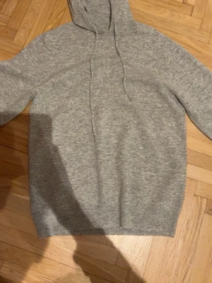 Grå kashmir hoodie  - Mysig grå stickad hoodie med huva och dragsnören. Tröjan har en enkel och clean design utan tryck eller logga, perfekt för dig som gillar en stilren look. Materialet ser ut att vara mjukt och lite tjockare, vilket gör den extra skön att bära.