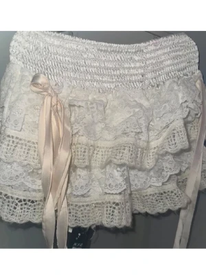 Designer Vit spetskjol med volanger och rosett - Supersöt kort vit kjol med flera lager spets och volanger. Kjolen har en bred smock i midjan och en sidenrosett på sidan som extra detalj. Perfekt för dig som gillar romantisk och feminin stil.    Aldrig använd. 