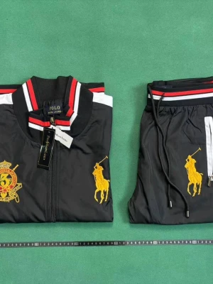 Svart set från Polo Ralph Lauren - Snyggt svart set från Polo Ralph Lauren med jacka och byxor. Setet har vita och röda detaljer, dragkedja, broderad gul logga och emblem på jackan samt byxorna. Byxorna har dragsko i midjan och vita ränder längs sidorna. Perfekt för en sportig och trendig look.