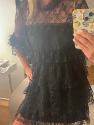Svart spetsklänning med volanger - Säljer en svart kort klänning i spets med volangdetaljer och transparenta partier. Klänningen har trekvartsärm och en romantisk look med flera lager spets. Perfekt för dig som vill ha en elegant och trendig outfit.