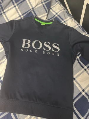Mörkblå sweatshirt från Hugo Boss, passar både S och M - Snygg mörkblå sweatshirt från Hugo Boss med stort BOSS-tryck i vitt framtill. Tröjan har rund halsringning, ribbade muddar och en grön detalj på insidan av kragen. Perfekt för en avslappnad och stilren look.