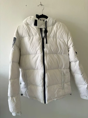 Vit pufferjacka med huva. Passar även L - Snygg vit pufferjacka med huva och svart dragkedja. Jackan har två sidofickor med dragkedja och en patch på vänster ärm. Justerbar huva med snörning och hög krage för extra värme. Perfekt för kalla dagar och riktigt stilren look.