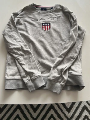 Grå sweatshirt från GANT - Snygg grå sweatshirt från GANT med broderad logga och ett sköldmärke med stjärnor och ränder på bröstet. Tröjan har rund halsringning, ribbade muddar vid ärmslut och nederkant samt en avslappnad passform. Perfekt för en casual look.