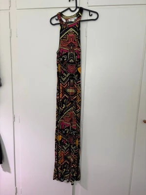 Mönstrad långklänning H&M Coachella - Färgstark långklänning från H&M Coachella-kollektionen med geometriskt mönster i svart, gul, orange och rosa. Klänningen har ärmlös design och rund halsringning. Perfekt för dig som vill sticka ut med en unik och bohemisk stil.