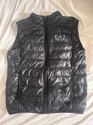 Svart dunväst från Emporio Armani - Snygg svart dunväst från Emporio Armani EA7 med quiltad design och hög krage. Västen har dragkedja framtill och logga på bröstet. Perfekt för lager-på-lager och ger en sportig vibe till din outfit.