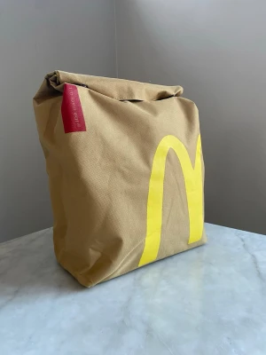 McDonald's ryggsäck - Stilren shoppingväska i beige tyg med McDonald's klassiska gula M-logga på framsidan och en röd etikett på sidan. Väskan har en mjuk, fyrkantig form och är tillverkad i kraftig bomull, perfekt för att bära hem dina grejer med extra attityd.