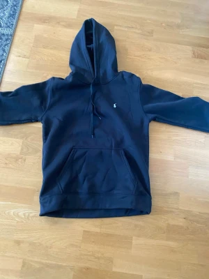 Svart hoodie från Polo Ralph Lauren - Klassisk svart hoodie från Polo Ralph Lauren med den ikoniska vita broderade loggan på bröstet. Hoodien har stor huva med dragsko och en rymlig magficka. Tillverkad i mjukt bomullsmaterial som är perfekt för chill dagar.