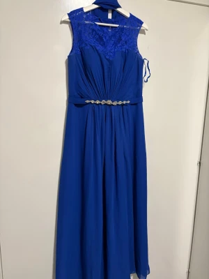 Blå långklänning från Romantica - Elegant blå långklänning från Romantica med spetsdetaljer upptill och ett vackert dekorerat bälte i midjan. Klänningen har ärmlös design och ett flytande, svepande tyg som ger en lyxig känsla. Perfekt för speciella tillfällen. Klänningen kommer med en långärmad tröja och en sjal ifall du känner ett behov av de.