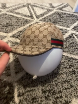 Gucci keps med monogram och ränder - Snygg beige keps från Gucci med klassiskt GG-monogram över hela kepsen och de ikoniska gröna och röda ränderna på sidan. Kepsen har böjd skärm och är tillverkad i canvasmaterial. Perfekt för dig som vill sticka ut med en lyxig accessoar.