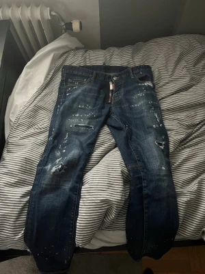 Mörkblå distressed jeans från Dsquared2 - Säljer ett par mörkblå jeans från Dsquared2 med coola slitningar, färgstänk och trasiga detaljer. Jeansen har klassisk femficksmodell, dragkedja och synliga sömmar. Perfekta för dig som gillar en edgy streetstil.