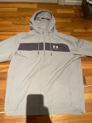 Ljusgrå vindjacka från Under Armour - Snygg ljusgrå vindjacka från Under Armour med dragkedja framtill, huva med dragsko och en mörkgrå panel över bröstet där loggan sitter. Jackan har två sidofickor och är tillverkad i ett lätt, vindtätt material som passar perfekt för blåsiga dagar.