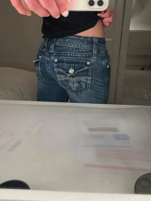 Rock revival jeans - Säljer ett par blå Rock Revival jeans med snyggt broderi och dekorativa knappar på bakfickorna. Jeansen har låg midja, raka ben och slitna detaljer för en cool look. Materialet är klassisk denim med stretch för skön passform.