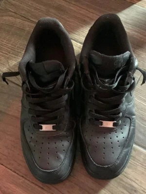 Svarta sneakers i läderstil - Snygga helsvarta sneakers från Nike med klassisk låg profil och perforerad tåbox. Skorna har svarta skosnören, metall-detalj på snörningen och en vadderad plös. Perfekta för en clean och stilren look.