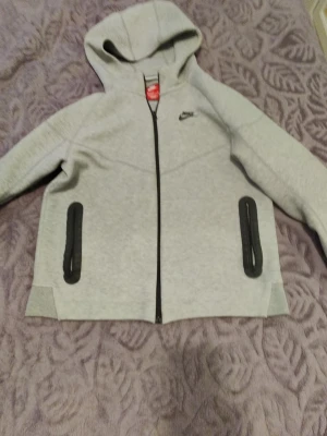 Grå zip hoodie från Nike - Snygg ljusgrå hoodie från Nike med dragkedja framtill och två svarta fickor med dragkedja. Klassisk huva och liten Nike-logga på bröstet. Perfekt för en sportig och avslappnad stil.