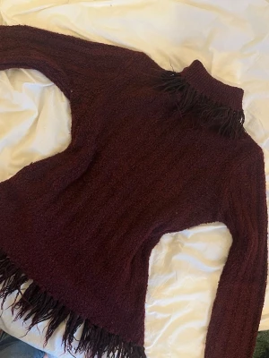 Vinröd stickad polotröja med fransar - Säljer en vinröd stickad polotröja med långa ärmar och coola fransdetaljer vid hals, ärmslut och nederkant. Tröjan har en figurnära passform och är gjord i ett mjukt, tjockt material. Skriv vid frågor och pris kan diskuteras💕💕