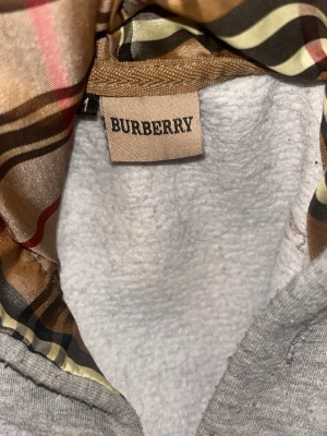 Grå zip hoodie från Burberry - Snygg grå hoodie från Burberry med dragkedja och klassiskt rutigt foder i huvan. Hoodien har en broderad logga på bröstet och är tillverkad i mjukt material som känns lyxigt. Perfekt för dig som vill ha en stilren och exklusiv look.