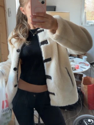 Beige teddyjacka med mörkbruka detaljer - Supermjuk och fluffig teddyjacka i beige med mörkbruna knappar och mörkbruna detaljer längs knäppningen och fickorna. Enkel att styla till både jeans och kjol.