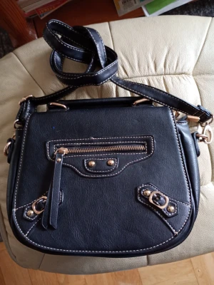 Svart axelväska med gulddetaljer - Snygg svart axelväska i konstläder med dekorativa guldfärgade nitar och dragkedjor. Väskan har justerbar axelrem, ficka fram med dragkedja och coola detaljer. Perfekt för dig som vill ha en stilren men edgy look.