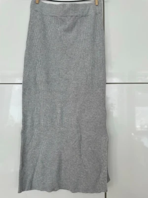 Grå ribbad långkjol med slits - Säljer en snygg grå långkjol i ribbat material. Kjolen har en stilren design med en slits på sidan som ger extra edge. Perfekt att matcha med en enkel topp eller hoodie för en chill look. Kjolen är lite sliten längs sömmen vid midjan (se på bild 4).