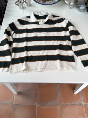 Randig rugbytröja med krage - Snygg långärmad rugbytröja med breda mörkgröna och vita ränder samt tunna bruna linjer. Klassisk vit krage och avslappnad passform. Perfekt för en sportig och trendig look.
