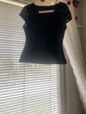 Svart topp med spetsdetaljer - Snygg svart topp med spets på axlarna och överdelen. Modellen är figurnära och har korta ärmar i spets som ger en elegant touch. Perfekt att styla med både jeans och kjol för en trendig look.