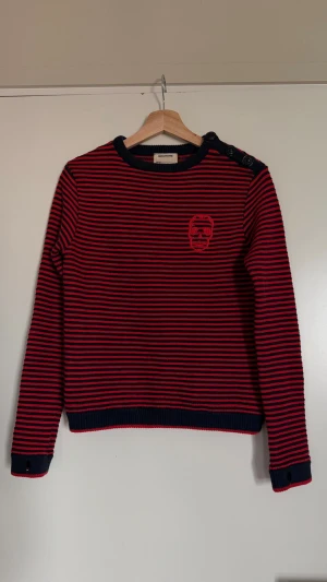 Randig tröja från Zadig & Voltaire - Säljer en cool röd och marinblå randig tröja från Zadig & Voltaire. Tröjan har rund hals, knappar på ena axeln och ett broderat emblem på bröstet. Tillverkad i mjukt stickat material och har ribbade muddar vid ärmslut och nederkant. Tröjan är i fint skick! 