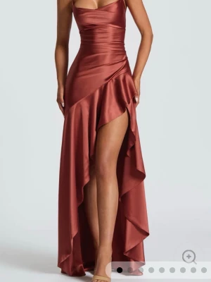Rostbrun bakklänning i satin - Snygg rostbrun långklänning i glansig satin med smala axelband och öppen rygg med snörning. Aldrig använd från VioletAurora! Klänningen har en asymmetrisk slits framtill och figurnära passform som framhäver siluetten. Perfekt för dig som vill sticka ut.