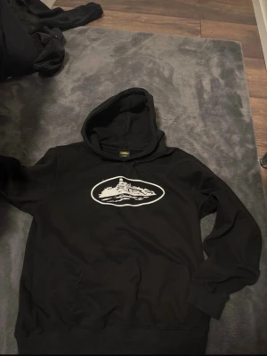 Svart hoodie med bergstryck - Säljer en svart hoodie med ett vitt tryck av ett berg i oval form på bröstet. Hoodien har en klassisk huva och en stor magficka framtill. Mjuk insida och ribbade muddar vid ärmar och nederkant. Perfekt för dig som gillar enkel och stilren streetwear.