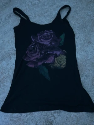 Svart linnetopp med rosor - Svart linnetopp med tryck av stora lila och gula rosor framtill. Tunn och lätt, perfekt för varma dagar. Smala axelband och figurnära passform. Snygg att styla till jeans eller kjol.