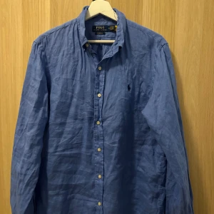 Blå linneskjorta från Polo Ralph Lauren - Snygg blå skjorta  från Polo Ralph Lauren med klassisk krage och knappar framtill. Skjortan har lång ärm och den ikoniska loggan broderad på bröstet. Perfekt för en stilren och avslappnad look. Tröjan ej strykt på bild, inga defekter och pris är absoult inte hugget, hör av er vid alla frågor!🌟