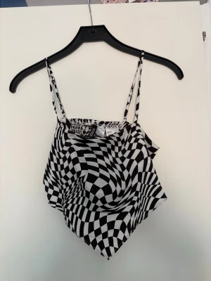 Svartvit mönstrad linnetopp - Trendig linnetopp med svartvitt optiskt rutmönster och smala axelband. Toppen har en bandana-liknande form och är tillverkad i ett lätt, slätt material. Perfekt för dig som vill sticka ut med en cool och modern look.