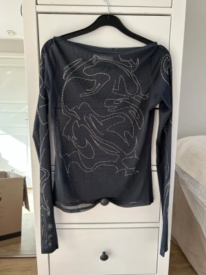 Svart mesh topp med strassdetaljer - Snygg svart mesh topp med långa ärmar och coola abstrakta mönster i strass över hela plagget. Toppen är genomskinlig och har en fin passform som ger en edgy vibe. Perfekt att styla lager på lager eller som statement piece. Några stenar har ramlat av.