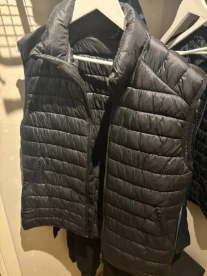 Svart quiltad dunväst - Snygg svart dunväst med quiltat mönster och hög krage. Västen har dragkedja framtill och två sidofickor med dragkedja. Perfekt att slänga över en hoodie eller tröja för extra värme. Enkel och stilren design som passar till det mesta.