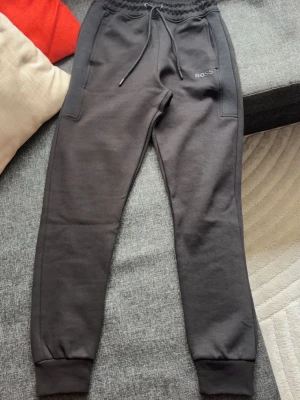 Svarta mjukisbyxor från BOSS - Säljer ett par svarta mjukisbyxor från BOSS med dragsko i midjan och ribbade muddar vid bensluten. Byxorna har två sidofickor och diskret BOSS-logga på vänster sida. Tillverkade i mjukt och bekvämt material, perfekta för chill dagar. Helt nya, bara haft på mig dom en gång men de var lite för tajta på mig. Skulle säga att dessa passar XS och S.
