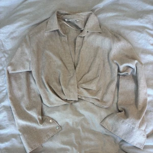Beige croppad topp från Zara - Säljer en beige croppad topp från Zara med twistad omlottdesign framtill. Blusen har lång ärm med bred manschett och klassisk krage. Det finns en dragkedja vid sidan! Använd en gång🥰