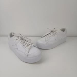 Nike Blazer Low Platform Vita Sneakers EUR 40 - Ett par vita sneakers med snörning. Skorna har en plattformssula och en diskret Nike-logotyp på sidorna. Hälen har också Nike-text. Skorna har en ren och klassisk design. Det finns en liten fläck på varje sko. 
