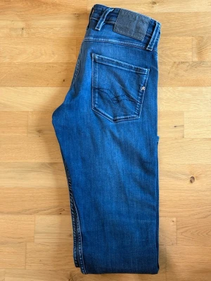 Blå slim fit jeans från Replay - Snygga blå slim fit jeans från Replay med klassisk femficksdesign och subtila slitningar framtill. Jeansen har en mörkblå tvätt och detaljerade sömmar, samt Replay-logga på bakfickan. Skicket är bra men dom är använda. Finns ett hål på sista bilden.