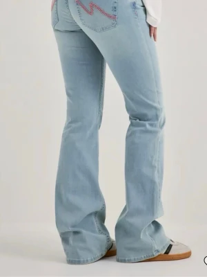 Ljusblå bootcut jeans med brodyr - Säljer ett par ljusblå bootcut jeans med rosa broderad detalj på bakfickan. Jeansen har hög midja och utsvängda ben, vilket ger en retroinspirerad look. Perfekta till sneakers eller boots för en avslappnad stil.