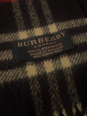 Burberry rutig halsduk i lammull - Klassisk halsduk från Burberry i 100% lammull. Mörkblå bas med vitt och rött rutmönster samt fransar i ändarna. Tillverkad i England och har den ikoniska Burberry-taggen. Perfekt accessoar för att lyfta din outfit.