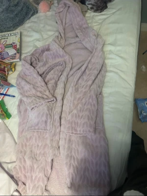 Ljuslila fluffig morgonrock med huva - Supermjuk och mysig ljuslila morgonrock med huva och två stora fickor framtill. Tillverkad i fluffigt fleece-material med diskret mönster. Perfekt för att chilla hemma och hålla sig varm under kalla dagar.