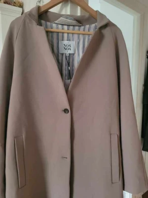 Beige kappa från Noa Noa, strl 46 - Stilren beige kappa från Noa Noa i storlek 46. Kappan har enkel knäppning framtill, två fickor och ett randigt innerfoder. Perfekt för höst och vinter med sin mjuka struktur och klassiska design.