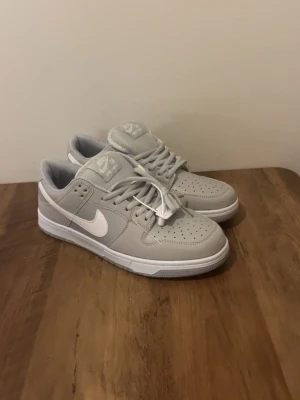 Nike SB grå Dunks - Snygga Nike SB sneakers i ljusgrått med vit swoosh och vita snören. Skorna har en klassisk låg siluett, perforerad tåbox och är tillverkade i en kombination av mocka och textil. Perfekta för dig som gillar stilrena och bekväma sneakers.