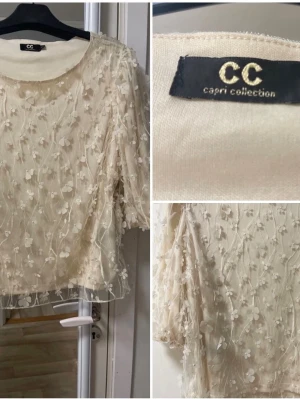 Krämvit blus med 3D-blommor Capri Collection - Romantisk krämvit blus från Capri Collection med transparent mesh och 3D-blommor över hela plagget. Blusen har rund halsringning och trekvartsärm. Perfekt för dig som gillar detaljer och en feminin look. 