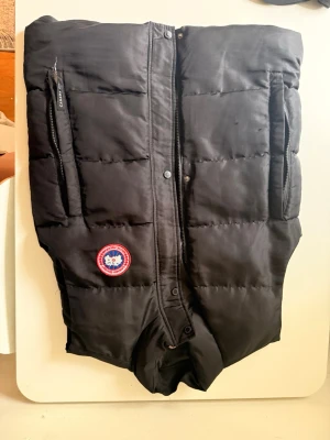 Svart dunväst från Canada Goose - Snygg svart dunväst från Canada Goose med klassisk logga på bröstet och diskret broderad etikett på axeln. Västen har två dragkedjeförsedda fickor framtill och quiltad design. Perfekt för lager-på-lager och håller dig varm under kyliga dagar.