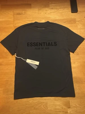 Svart Essentials Fear of God t-shirt - Snygg svart t-shirt från Essentials Fear of God med stor logga tryckt på bröstet. Klassisk passform och rund halsringning. Perfekt för dig som gillar streetwear och vill ha en stilren look.