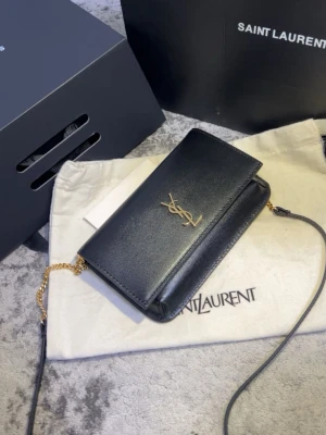 Ysl phone holder  - Saint Laurent Phone Holder köpt på Vinted under 2025 med Vinteds artikelverifiering. Produkten autentiserades och godkändes av Vinted innan den skickades till mig.  Det som medföljer:  * Originalbox * Dust bag * Original shoppingpåse * Digitalt originalkvitto från Saint Laurent * Vinteds digitala bekräftelse på att produkten verifierades och godkändes, Serienumret finns i väskan och kan visas för seriösa köpare vid intresse, Väskan är även behandlad med läderkräm,