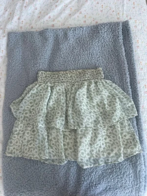 Blommig volangkjol - Supersöt kort kjol med volanger och blommigt mönster i ljusgrönt och vitt. Kjolen har en smockad midja som gör den bekväm och lätt att bära. Är i storlek 158/164 men passar mig som har xs. Kan finnas lösa trådar💗